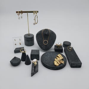 Peut inclure: Une collection de bijoux dorés, comprenant des boucles d'oreilles, des colliers, des bagues et une broche, exposés sur des présentoirs en pierre gris foncé et un présentoir à boucles d'oreilles en acrylique transparent. Les bijoux présentent des perles et des motifs circulaires.
