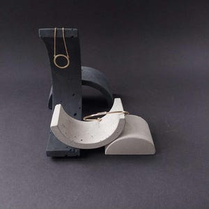 May include: A gold necklace with a circular pendant and a gold bracelet with small embellishments are displayed on gray and white concrete jewelry stands. The stands have a modern, minimalist design against a dark background.