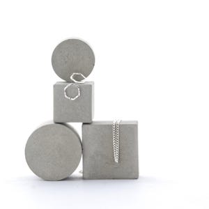 May include: Silver rings and a necklace displayed on grey concrete geometric shapes. The rings have textured designs. The necklace has a delicate chain. The background is white. The shapes include cylinders and cubes.