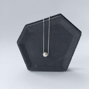 May include: A delicate silver chain necklace with a single, round, white pearl pendant. The necklace is displayed on a dark gray, geometric-shaped stand, creating a minimalist and elegant aesthetic. The pearl is approximately 0.3 inches in diameter.