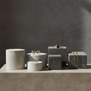 Könnte beinhalten: Präsentation von Goldschmuck, darunter eine Halskette, Ohrringe und Ringe, auf grauen Betonpodesten. Der Schmuck weist verschiedene Designs auf, darunter eine Anhängerhalskette, Creolen und ein Ring mit einem schwarzen Stein. Der Hintergrund ist ein neutrales Grau.