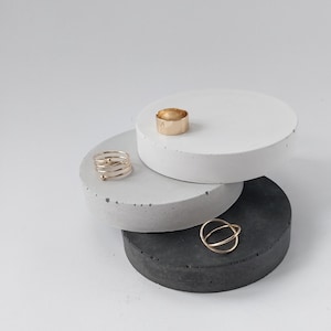 Peut inclure: Anneaux dorés présentés sur trois plateformes rondes en béton empilées. Les anneaux comprennent une bande épaisse, un design en spirale et un style à double bande. Les plateformes sont blanches, grises et noires, créant une esthétique minimaliste.