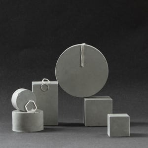 May include: A silver necklace hangs from a grey circular display in a studio shot. Geometric concrete shapes, including cylinders, cubes, and a rectangular block, are arranged to showcase the jewellery. The background is a dark grey.
