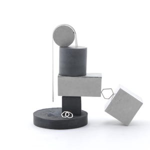 May include: An abstract sculpture made of grey concrete geometric shapes. A silver chain hangs from a circular piece, connected to a cylindrical form. A silver necklace and a hexagonal ring are also present.