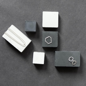 Peut inclure: Un assortiment de bijoux et de blocs d'exposition. Un collier en argent repose sur un bloc rectangulaire blanc. D'autres blocs sont gris et blancs, certains avec des boucles d'oreilles géométriques en argent. Le fond est gris foncé.