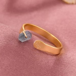Könnte beinhalten: Ein goldfarbener offener Ring mit einem hellblauen Edelstein. Der Ring ist vor einem rosa Satinhintergrund platziert, mit ein paar weißen Blumen in der oberen rechten Ecke. Das Design des Rings ist schlicht und elegant.
