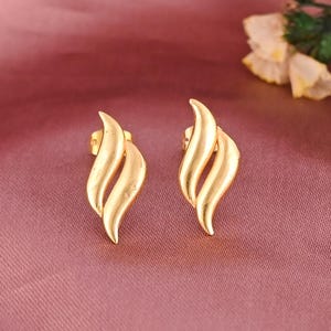 May include: A pair of gold-coloured stud earrings. Each earring has a flame-like design with curved lines. The earrings are set against a pink background, with white flowers in the upper right corner.