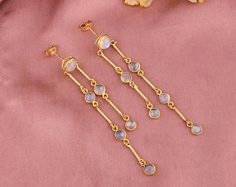 May include: Gold-toned dangle earrings featuring a series of small, round, pale blue gemstones. The earrings have a linear design with a post-back closure. The background is a solid, muted pink.
