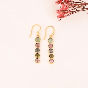 May include: A pair of gold-coloured drop earrings. Each earring has a vertical row of six round, multi-coloured gemstones. The gemstones include shades of green, pink, and grey. The earrings are displayed on a white background.