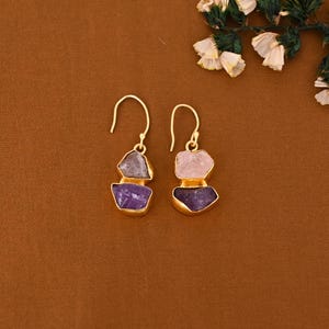 Könnte beinhalten: Ein Paar goldfarbene Ohrringe mit rohen Amethyst- und Rosenquarz-Edelsteinen. Die Ohrringe haben ein geschichtetes Design mit einem goldfarbenen Haken. Die Edelsteine sind lila, grau und rosa.
