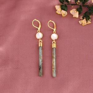 Puede incluir: Un par de pendientes colgantes dorados. Cada pendiente presenta una piedra blanca y circular en la parte superior, conectada a una piedra larga, rectangular, gris y verde. Los pendientes se muestran sobre un fondo rosa, con detalles florales.