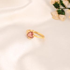 Op de afbeelding: Een gouden ring met een driehoekige, roze edelsteen. De ring ligt op een witte, zijdeachtige ondergrond, met gedroogde bloemen op de achtergrond. De ring heeft een dunne, delicate band.