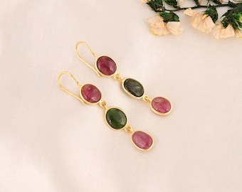 May include: Gold-tone earrings with a drop design. Each earring features three oval-shaped gemstones: a deep red, a dark green, and a lighter red. The earrings are displayed on a white surface, with floral accents in the background.