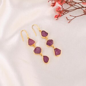 Peut inclure: Boucles d'oreilles dorées avec trois pierres précieuses roses foncées brutes et de forme irrégulière suspendues à un crochet. Les boucles d'oreilles sont présentées sur une surface blanche et soyeuse, avec une branche de fleurs roses en arrière-plan.