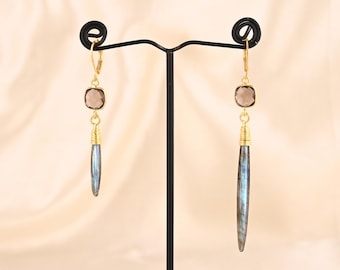 May include: A pair of gold-colored earrings with a square, smoky quartz stone at the top and a long, blue-gray labradorite stone pendant. The earrings are displayed on a black jewelry stand against a cream-colored background.