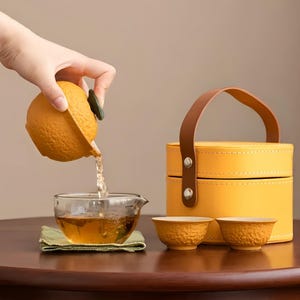 以下が含まれることがあります： 緑色の葉の取っ手が付いたオレンジ型の陶器製ティーポットが、透明なガラスのカップに茶を注いでいます。その横には、オレンジ色の質感を持つ陶器製の茶碗2つと、茶色のレザーハンドルが付いた黄色の円筒形キャリングケースが置かれています。