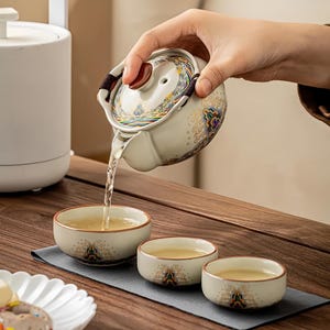 May include: A ceramic tea set with a teapot pouring tea into three small cups. The teapot and cups are cream-colored with colorful decorative patterns. The tea is a light amber color. The set is on a wooden surface.