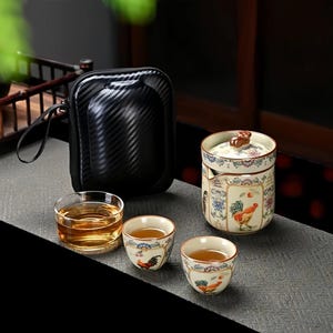 Puede incluir: Un juego de té con un estuche negro, un recipiente de té de cerámica con un diseño de gallo, dos tazas de té pequeñas con ilustraciones de gallos y una taza de té de vidrio transparente llena de té. El juego está dispuesto sobre una superficie gris.