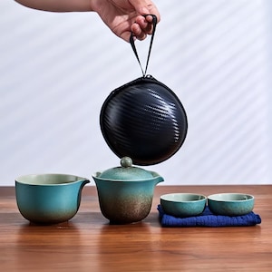 Pode incluir: Um conjunto de chá com acabamento esmaltado turquesa e castanho. O conjunto inclui uma chaleira com tampa, duas chávenas pequenas e uma chávena maior. Uma mala de transporte preta é segurada acima do conjunto. O conjunto de chá está numa superfície de madeira.