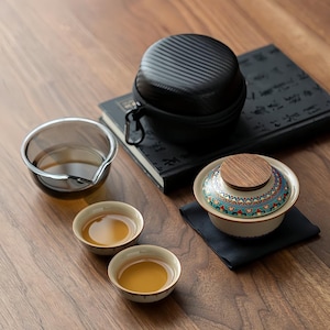 May include: A tea set with a black carrying case, a glass pitcher, two small tea cups, and a decorative tea bowl with a wooden lid. The tea bowl has a colorful patterned design. The items are arranged on a wooden surface.