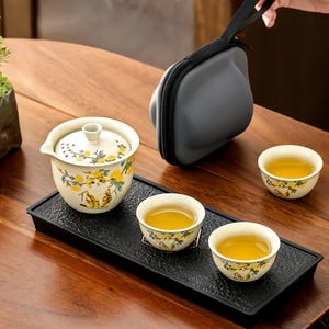 Puede incluir: Un juego de té con una tetera crema y tres tazas a juego, todas decoradas con diseños florales amarillos y de tigre. El juego incluye una bandeja negra y un estuche de transporte gris. Las tazas están llenas de té.
