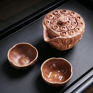 Puede incluir: Una tetera de cerámica marrón con una tapa texturizada que se asemeja a una vaina de semillas de loto y dos tazas de té pequeñas, redondas y acanaladas a juego. La tetera presenta un pico y una textura de superficie orgánica detallada.