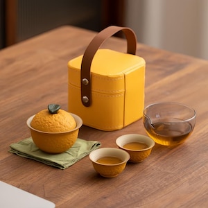 May include: A yellow tea set with a matching carrying case. The set includes a textured tea bowl with a lid, two small cups, and a clear glass pitcher. The tea is amber-coloured, and the case has a brown handle.