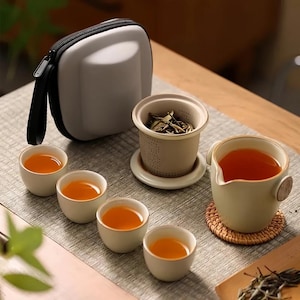 Op de afbeelding: Een theeset met een lichtgrijze draagtas, kleine witte theekopjes met gouden randen, een thee-ei en een theepot. De theekopjes en de pot zijn gevuld met thee. De set is op een lichtgrijze ondergrond geplaatst.
