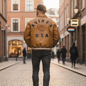 K&ouml;nnte beinhalten: Hellbraune Lederjacke mit dem Text "AVIREX USA" auf dem R&uuml;cken. Die Jacke hat einen braun-wei&szlig; gestreiften Saum und B&uuml;ndchen. Die Person tr&auml;gt eine dunkelgraue Jeans und schwarz-wei&szlig;e Turnschuhe.