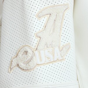 Puede incluir: Primer plano de una chaqueta de cuero blanco perforada con un parche color crema. El parche presenta la letra "J", las letras "USA" y un dise&ntilde;o estilizado. La chaqueta tiene una est&eacute;tica deportiva.
