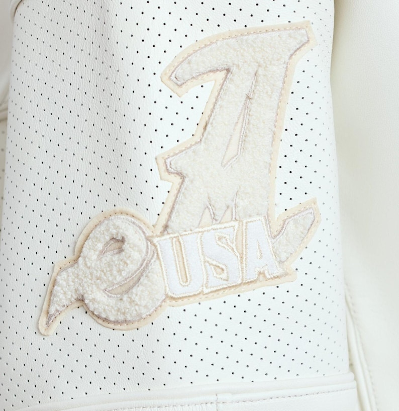 Puede incluir: Primer plano de una chaqueta de cuero blanco perforada con un parche color crema. El parche presenta la letra "J", las letras "USA" y un dise&ntilde;o estilizado. La chaqueta tiene una est&eacute;tica deportiva.