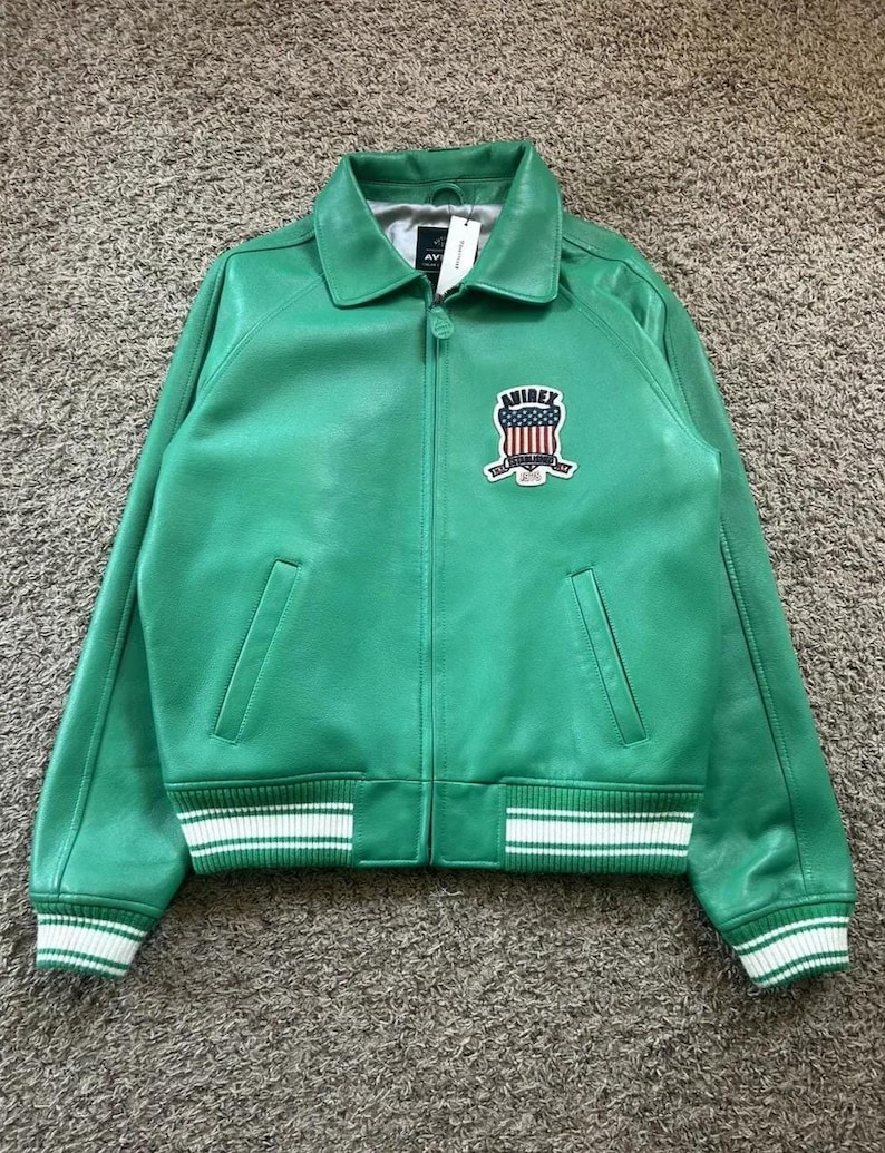 Puede incluir: Chaqueta de cuero verde de estilo bomber cl&aacute;sico. Cuello camisero, cremallera frontal y dos bolsillos delanteros. Los pu&ntilde;os y el dobladillo son a rayas blancas y verdes. Un parche bordado con el texto "AVIREX" y un dise&ntilde;o patri&oacute;tico est&aacute; en el pecho.