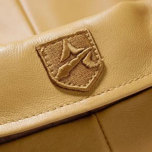 K&ouml;nnte beinhalten: Nahaufnahme eines Details einer hellbraunen Lederjacke. Die Jacke hat ein aufgesticktes Emblem auf der oberen Brust. Das Emblem hat die Form eines Schildes mit einem detaillierten, gestickten Design in passender hellbrauner Farbe. Das Leder hat eine glatte Textur.