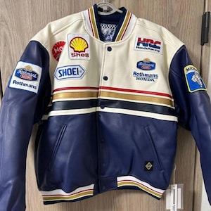 以下が含まれることがあります： シェル、ショウエイ、ロスマンズなどのスポンサーパッチが付いた、青とクリーム色のレーシングジャケット。 ジャケットはスナップボタン留め、青い袖、胸にストライプのデザインが施されています。 ジャケットはハンガーに吊るされています。