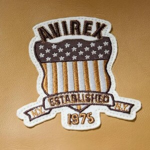 K&ouml;nnte beinhalten: Gestickter Aufn&auml;her mit dem Text "AVIREX" &uuml;ber einem Schilddesign mit Sternen und Streifen sowie dem Text "ESTABLISHED 1975". Der Aufn&auml;her hat einen wei&szlig;en Rand und ist auf einem beigefarbenen Hintergrund platziert.