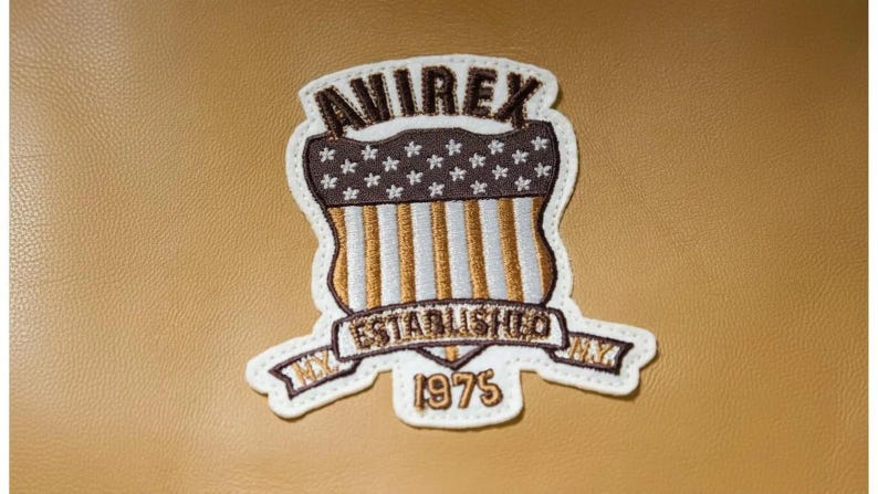 K&ouml;nnte beinhalten: Gestickter Aufn&auml;her mit dem Text "AVIREX" &uuml;ber einem Schilddesign mit Sternen und Streifen sowie dem Text "ESTABLISHED 1975". Der Aufn&auml;her hat einen wei&szlig;en Rand und ist auf einem beigefarbenen Hintergrund platziert.