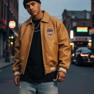 K&ouml;nnte beinhalten: Hellbraune Lederjacke mit Kragen, schwarzes T-Shirt und hellblaue Jeans. Die Jacke hat einen Aufn&auml;her auf der Brust und wei&szlig;-beige gestreifte Manschetten. Die Person tr&auml;gt eine schwarze M&uuml;tze und eine silberne Kette.