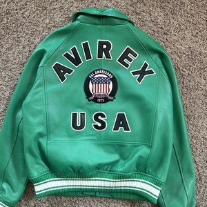 Puede incluir: Chaqueta de cuero verde con detalles en blanco y negro. La parte trasera presenta el texto "AVIREX" y "USA" en letras blancas. Tambi&eacute;n se ve un parche circular con la bandera estadounidense. La chaqueta tiene dobladillo y pu&ntilde;os acanalados.
