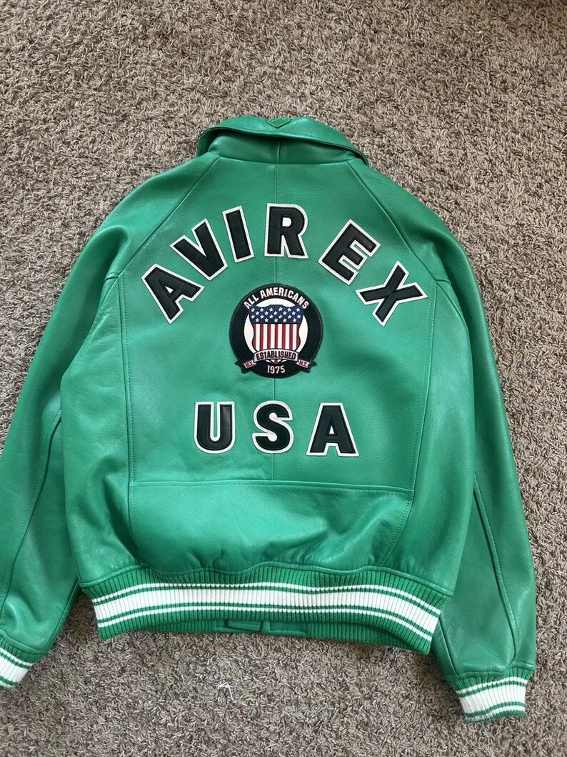 Puede incluir: Chaqueta de cuero verde con detalles en blanco y negro. La parte trasera presenta el texto "AVIREX" y "USA" en letras blancas. Tambi&eacute;n se ve un parche circular con la bandera estadounidense. La chaqueta tiene dobladillo y pu&ntilde;os acanalados.
