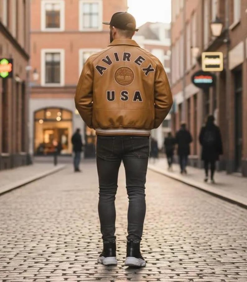 K&ouml;nnte beinhalten: Hellbraune Lederjacke mit dem Text "AVIREX USA" auf dem R&uuml;cken. Die Jacke hat einen braun-wei&szlig; gestreiften Saum und B&uuml;ndchen. Die Person tr&auml;gt eine dunkelgraue Jeans und schwarz-wei&szlig;e Turnschuhe.