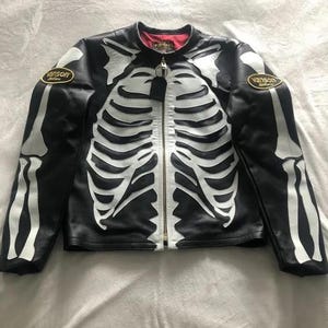 Könnte beinhalten: Schwarze Lederjacke mit weißem Skelettmuster. Die Jacke hat einen durchgehenden Reißverschluss, ein rotes Innenfutter und "Vanson" Aufnäher auf den Ärmeln. Ein einzigartiges und auffälliges Kleidungsstück.