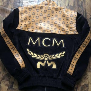 May include: Black velvet jacket with gold MCM logo and accents. The jacket features a tan and gold patterned yoke and sleeve stripes with the MCM logo. The jacket has a full zipper and ribbed cuffs and hem.