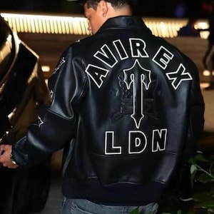 Könnte beinhalten: Schwarze Lederjacke mit weißen Buchstaben, die "AVIREX" und "LDN" lesen. Auf der Rückseite der Jacke befindet sich ein großes Emblem. Die Person trägt eine schwarze Kappe und eine graue Jeans.