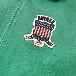 Puede incluir: Primer plano de una chaqueta de cuero verde brillante con un parche. El parche presenta la palabra "AVIREX" sobre un dise&ntilde;o de escudo con rayas rojas y blancas y estrellas azules. El parche tambi&eacute;n incluye el texto "NY ESTABLISHED" y el a&ntilde;o "1975".