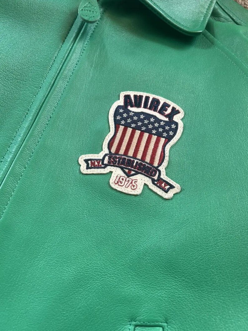 Puede incluir: Primer plano de una chaqueta de cuero verde brillante con un parche. El parche presenta la palabra "AVIREX" sobre un dise&ntilde;o de escudo con rayas rojas y blancas y estrellas azules. El parche tambi&eacute;n incluye el texto "NY ESTABLISHED" y el a&ntilde;o "1975".