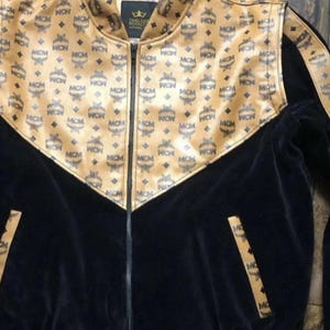 May include: A black and tan zip-up jacket with a repeating MCM logo pattern. The jacket features a black velvet body and tan accents on the shoulders and collar. The jacket has a high collar and a zipper closure.