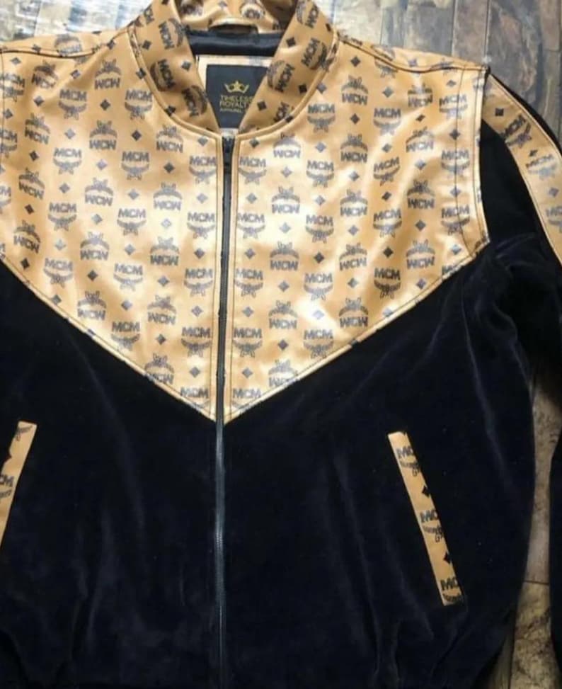 May include: A black and tan zip-up jacket with a repeating MCM logo pattern. The jacket features a black velvet body and tan accents on the shoulders and collar. The jacket has a high collar and a zipper closure.