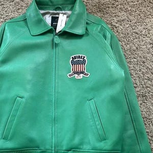 Puede incluir: Chaqueta de cuero verde brillante con cuello, cremallera y dos bolsillos delanteros. La chaqueta presenta un parche con un dise&ntilde;o rojo, blanco y azul y el texto "AVIREX ESTABLISHED 1975". Rayas blancas adornan los pu&ntilde;os y el dobladillo.