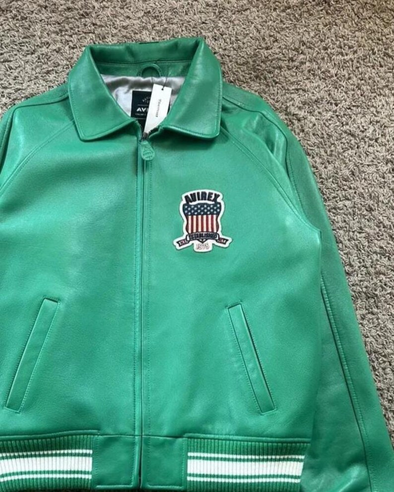 Puede incluir: Chaqueta de cuero verde brillante con cuello, cremallera y dos bolsillos delanteros. La chaqueta presenta un parche con un dise&ntilde;o rojo, blanco y azul y el texto "AVIREX ESTABLISHED 1975". Rayas blancas adornan los pu&ntilde;os y el dobladillo.