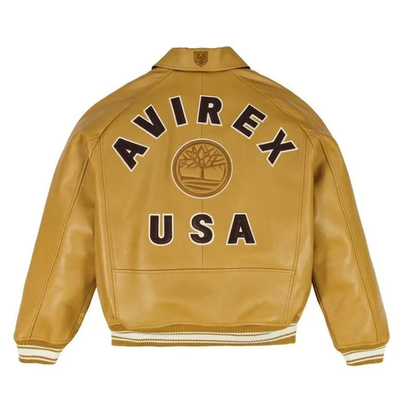 K&ouml;nnte beinhalten: Hellbraune Lederjacke mit dem Wort "AVIREX" in braunen Buchstaben auf dem oberen R&uuml;cken. Darunter befindet sich ein rundes Emblem mit einem Baumdesign und dem Wort "USA". Die Jacke hat einen Kragen und gerippte B&uuml;ndchen.